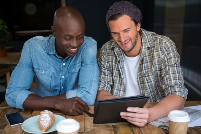 Two male friends are sitting at a table in a coffee shop, smiling and using a digital tablet. They are casually dressed and appear to be enjoying their time together. This image can be used to depict friendship, modern technology, and leisure activities. It is suitable for use in advertisements, blogs, and social media posts related to lifestyle, technology, and social interactions.