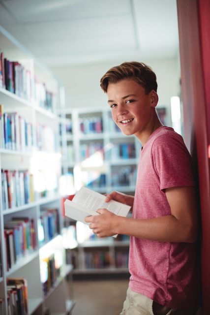 Teenage boy smiling while holding a book in a school library. Ideal for educational materials, school promotions, and library campaigns. Perfect for content related to reading, studying, and academic success.