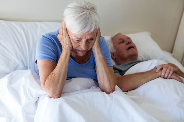 Senior woman sitting up in bed, covering her ears, looking frustrated while her partner is snoring beside her. This image can be used to depict sleep disturbances, relationship challenges, health issues related to aging, and the impact of snoring on sleep quality. Ideal for articles on sleep health, elderly care, and relationship advice.