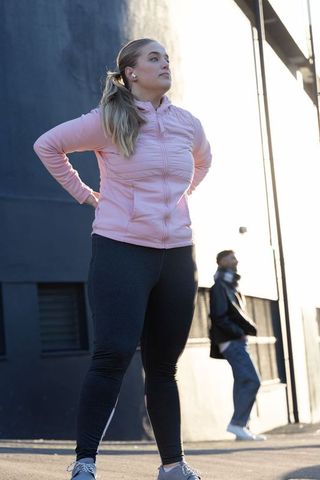 Woman standing hands on hips in pink jacket during city warmup, urban fitness portrait
