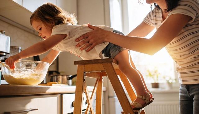 Toddler reaching and stirring batter as parent steadying on wooden stool in sunlit kitchen