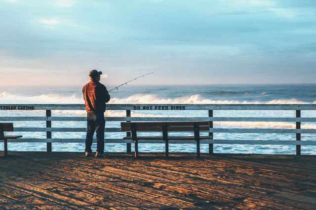 Solitary fisherman standing on ocean pier, casting fishing line into the water as the sun rises. Ideal for concepts related to outdoor activities, relaxation, hobbies, and enjoying nature's tranquility.