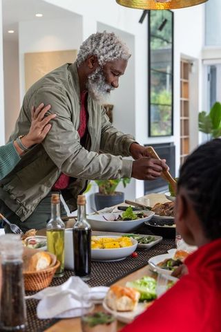 African american senior man serving family dinner around sunlit table in modern kitchen
