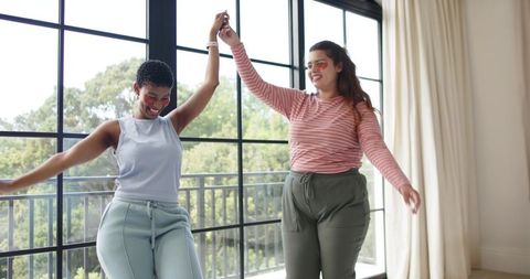 Two friends dancing together in sunlit lounge by black-framed windows and balcony