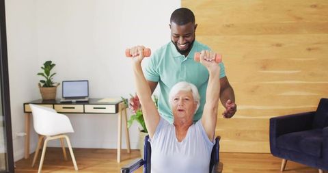 Senior woman exercising with caregiver guiding overhead dumbbell lifts in wheelchair
