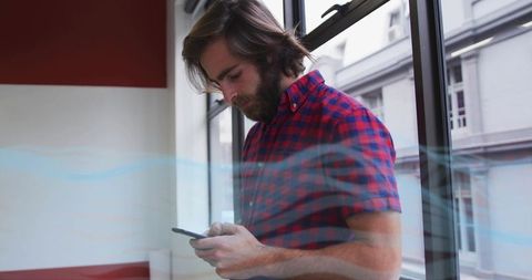 Bearded man checking smartphone by window in casual red plaid shirt with urban reflections
