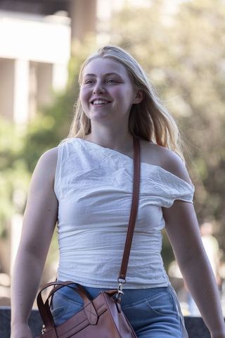 Young woman walking in sunny urban street smiling in white one-shoulder top carrying leather bag