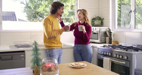 Diverse couple sharing cozy kitchen moment, smiling with mugs and cookies in warm sunlight