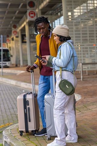 Diverse young travelers checking smartphone at bus platform with suitcases wearing beanie