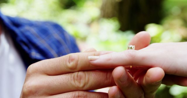A close-up captures a young man placing an engagement ring on a young woman's finger, symbolizing a marriage proposal. Set against a blurred natural backdrop, the moment signifies a commitment to a shared future.