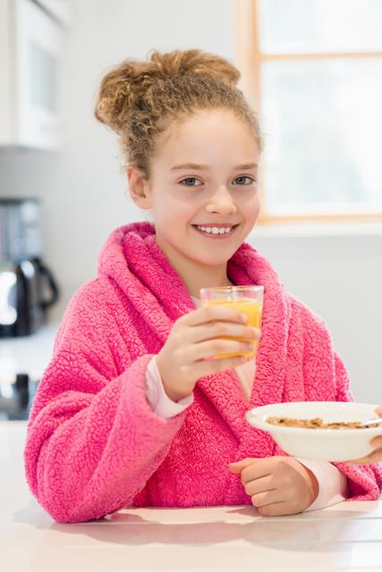 Young girl enjoying breakfast in a bright kitchen, wearing a cozy pink robe. She is holding a glass of orange juice and a bowl of cereal, smiling at the camera. Perfect for use in advertisements for healthy eating, morning routines, family lifestyle, or home comfort products.