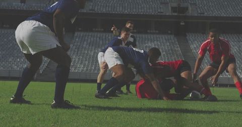 Rugby players contesting ruck on stadium pitch, navy and red jerseys, referee overseeing