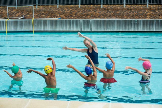 Female instructor teaching children swimming techniques in a pool. Children wearing colorful swim caps and swimwear, practicing arm movements. Ideal for use in educational materials, sports and fitness promotions, summer camp advertisements, and health and wellness campaigns.