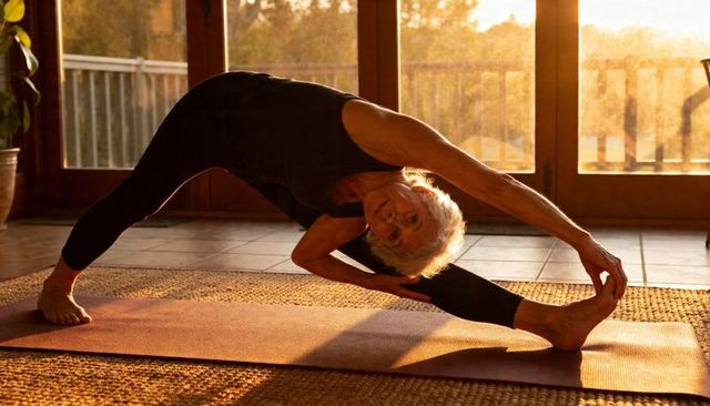 Senior woman practicing deep side stretch on yoga mat in sunlit home at golden hour