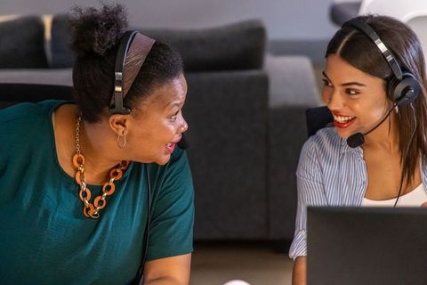 Diverse female coworkers chatting and collaborating with headsets and laptop in modern office lounge