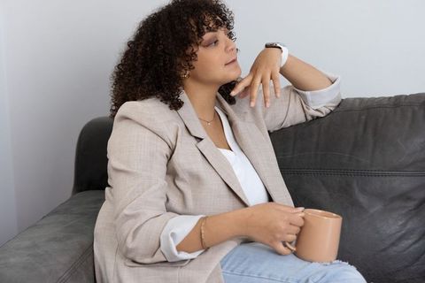 African american woman lounging on sofa holding mug wearing blazer and smartwatch