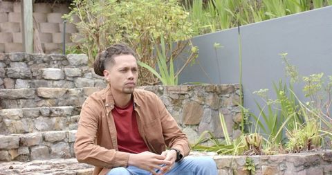 Contemplative man sitting on stone steps in terraced garden wearing brown jacket
