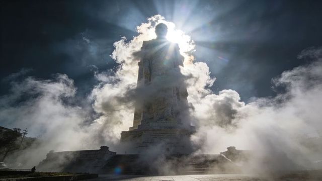 Backlit monument emerging through mist and sunburst on urban plaza, dramatic silhouette