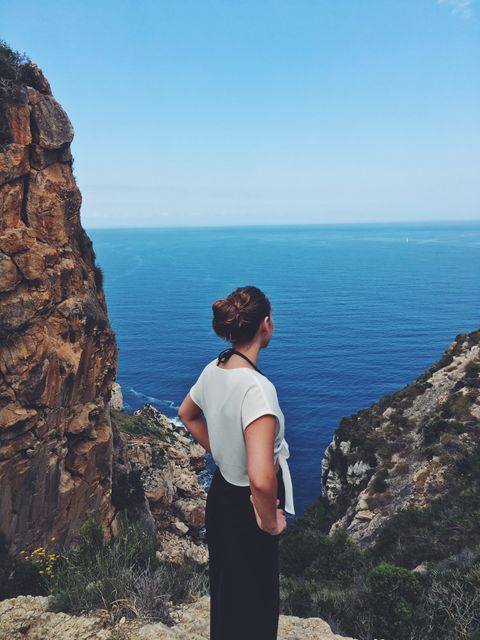 A woman stands on a cliffside, looking at the expansive ocean view. It is ideal for travel brochures, adventure magazines, nature blogs, and promoting outdoor activities. The scene exudes a sense of tranquility and the grandeur of nature, making it perfect for campaigns related to mental well-being, exploration, and relaxation.