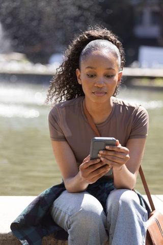 African american woman checking smartphone by park fountain in sunlight
