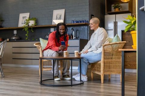 Diverse friends sharing smartphone and coffee in cozy modern kitchen nook, collaborating