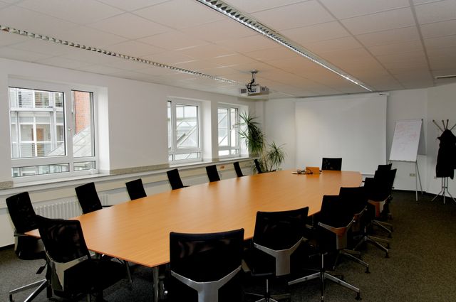Spacious conference room equipped with a long wooden table, black office chairs, and a projector. The room features large windows allowing natural light. This setting is perfect for business meetings, corporate gatherings, presentations, and professional seminars. Useful for business-related content, office environment illustrations, and corporate presentations.