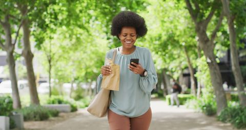 Happy Woman Strolling in Park with Smartphone and Brown Bag