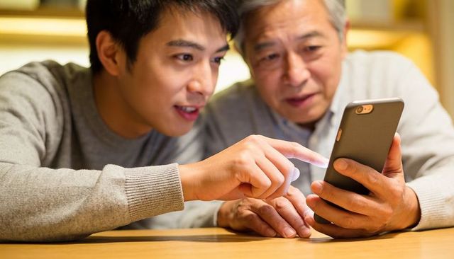 Adult son teaching senior father smartphone skills at cozy wooden table, sharing technology