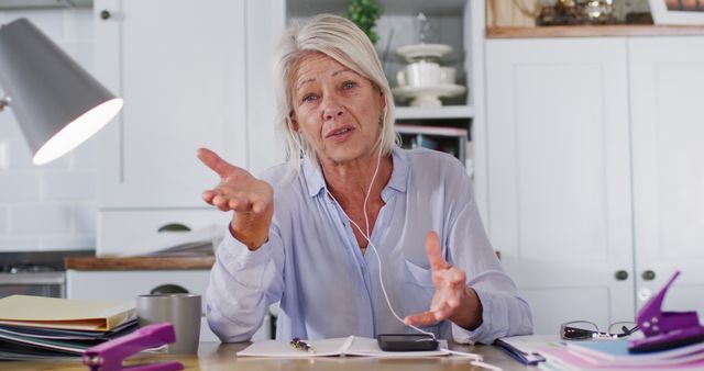 Senior woman engaging in a video call from her home office. She is speaking and gesturing with her hands while wearing headphones. Papers and documents are spread out on the desk, signifying a work environment. Ideal for themes related to remote working, online communication for seniors, technologically savvy elderly individuals, and home office setups.