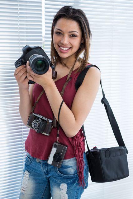 Young female photographer smiling while holding a camera in a modern studio. She is dressed casually and has multiple cameras and a camera bag. Ideal for use in articles about photography, creative professions, or hobbyist photographers.