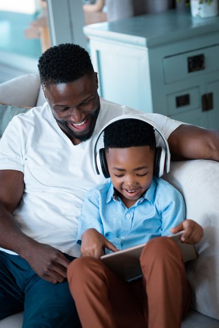 African American father and son sitting on a sofa, enjoying time together while using a tablet. The son is wearing headphones and smiling, indicating a fun and engaging activity. This image is perfect for illustrating family bonding, modern parenting, and the use of technology in everyday life. Ideal for use in advertisements, blogs, and articles about family life, technology, and home entertainment.