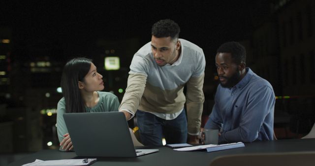 Image of three diverse male and female colleagues in discussion around a laptop at night in office. Business, communication, inclusivity and flexible working concept digitally generated image.