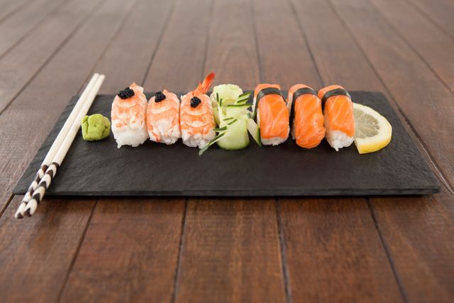 Nigiri sushi arranged on black stone slate with chopsticks on wooden table. Includes salmon and shrimp nigiri, wasabi, cucumber slices, and lemon slice. Ideal for use in food blogs, restaurant menus, culinary websites, and Japanese cuisine promotions.