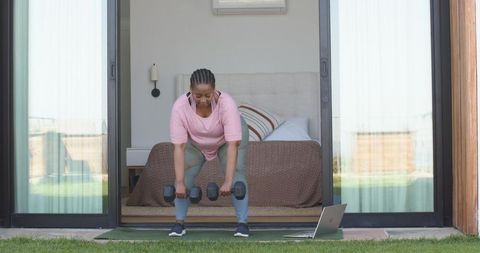 African american woman working out in doorway lifting dumbbells with laptop
