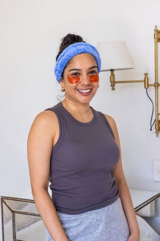 Smiling woman wearing blue plush headband and orange under-eye patches in cozy bedroom