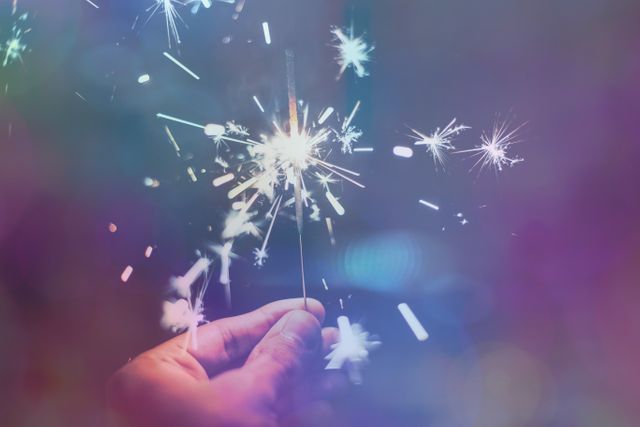 Close up of hand holding a burning sparkler against bokeh spots of light in background. Festivity and celebration concept