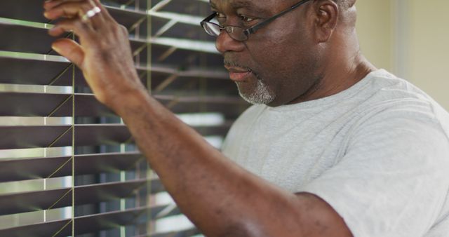 Senior man wearing eyeglasses and casual clothing adjusting window blinds. He appears thoughtful while looking through the blinds, possibly anticipating or observing something. Suitable for illustrating themes like home management, daily routines, senior lifestyle, and moments of reflection.