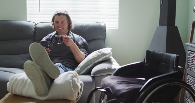 Relaxed Man in Living Room Enjoying Coffee Holding Smartphone With Wheelchair by Couch - Download Free Stock Images Pikwizard.com