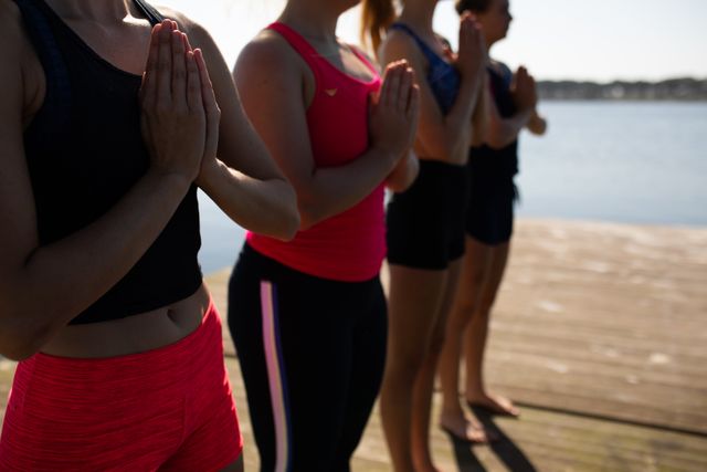 Side view mid section of a rowing team of four Caucasian women wearing sportswear and training on a jetty by the river, standing in a row, practicing yoga and meditating with river and blue sky in the background.