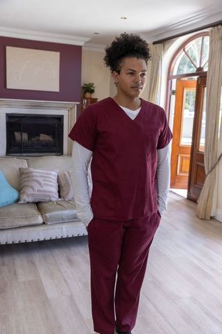 Caregiver wearing maroon scrubs standing hands in pockets near arched door and fireplace