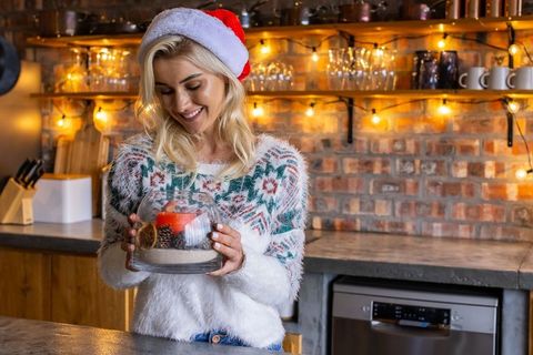 Blonde woman in santa hat arranging holiday candle in glass cloche on rustic kitchen counter