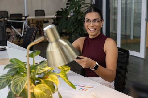 Young female professional laughing while using smartphone at bright open-plan office desk