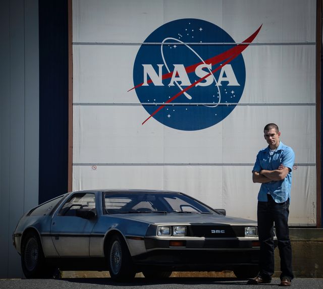 Man standing next to DeLorean car in front of NASA Goddard Space Flight Center sign Greenbelt, Maryland. Ideal for content about science fiction, classic cars, NASA, movie references, technology predictions, automotive history. Could be used in blogs about 'Back to the Future', articles on DeLorean cars, or NASA's involvement in popular culture.