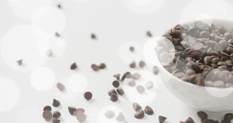 Minimalist high-key still life of dark chocolate chips spilling from white bowl with bokeh