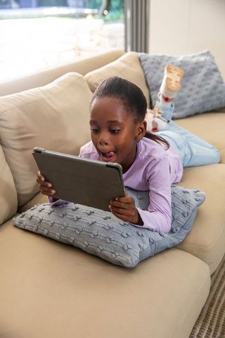 African american girl lying on sofa using tablet, enjoying screen time in living room
