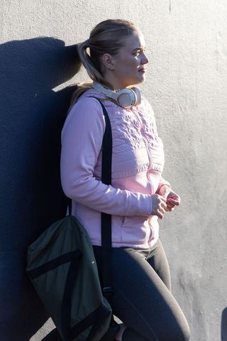 Young woman leaning on sunlit wall wearing pink hoodie and headphones