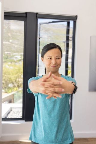 Asian mature woman stretching arms in sunlit home by black-framed windows wearing teal shirt smartwa