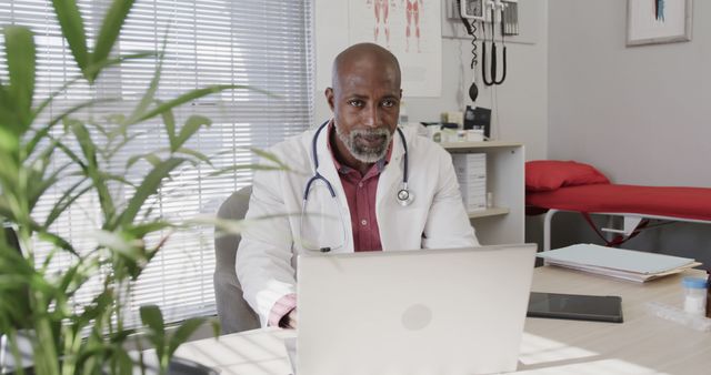 Senior male doctor with beard working on laptop in a medical office. Background depicts a modern, well-equipped medical environment, including anatomical charts and medical records. Use this image for promoting healthcare services, telemedicine solutions, healthcare blog posts, or informational content on medical websites.