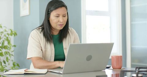 Asian woman working on laptop in bright home office, focused remote work and productivity