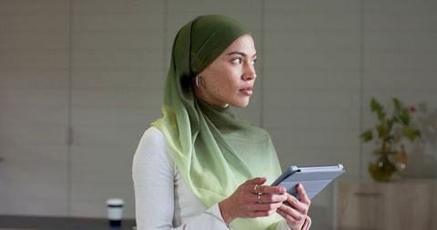 Indian woman wearing green hijab holding tablet looking right in modern office